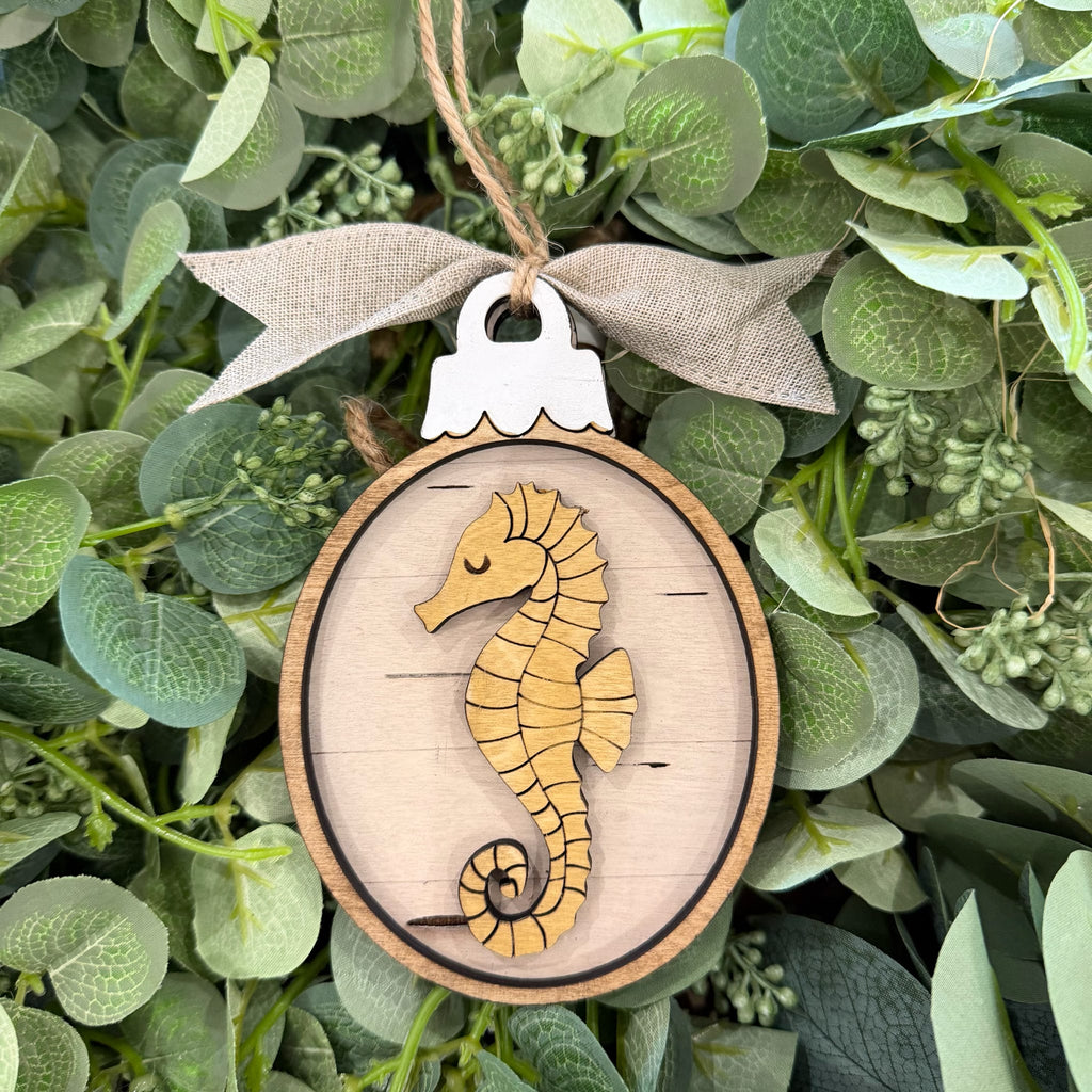 A wooden seahorse ornament hanging from a burlap ribbon, set against a background of green eucalyptus leaves.