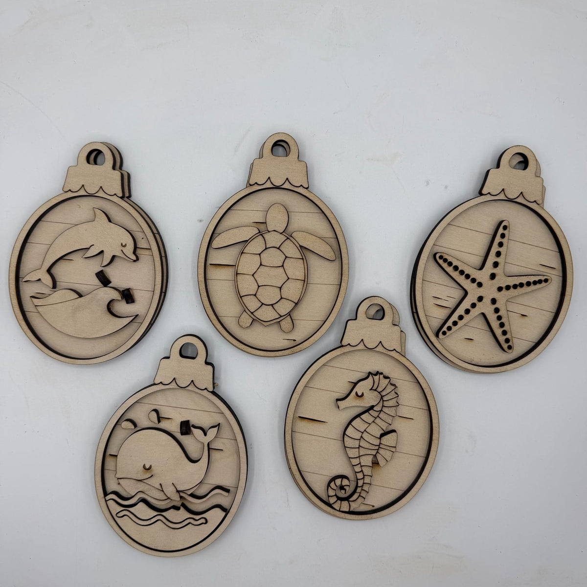 Five oval unfinished wood ornaments with sea-themed engravings, including a dolphin, turtle, starfish, whale, and seahorse, displayed on a light gray background.