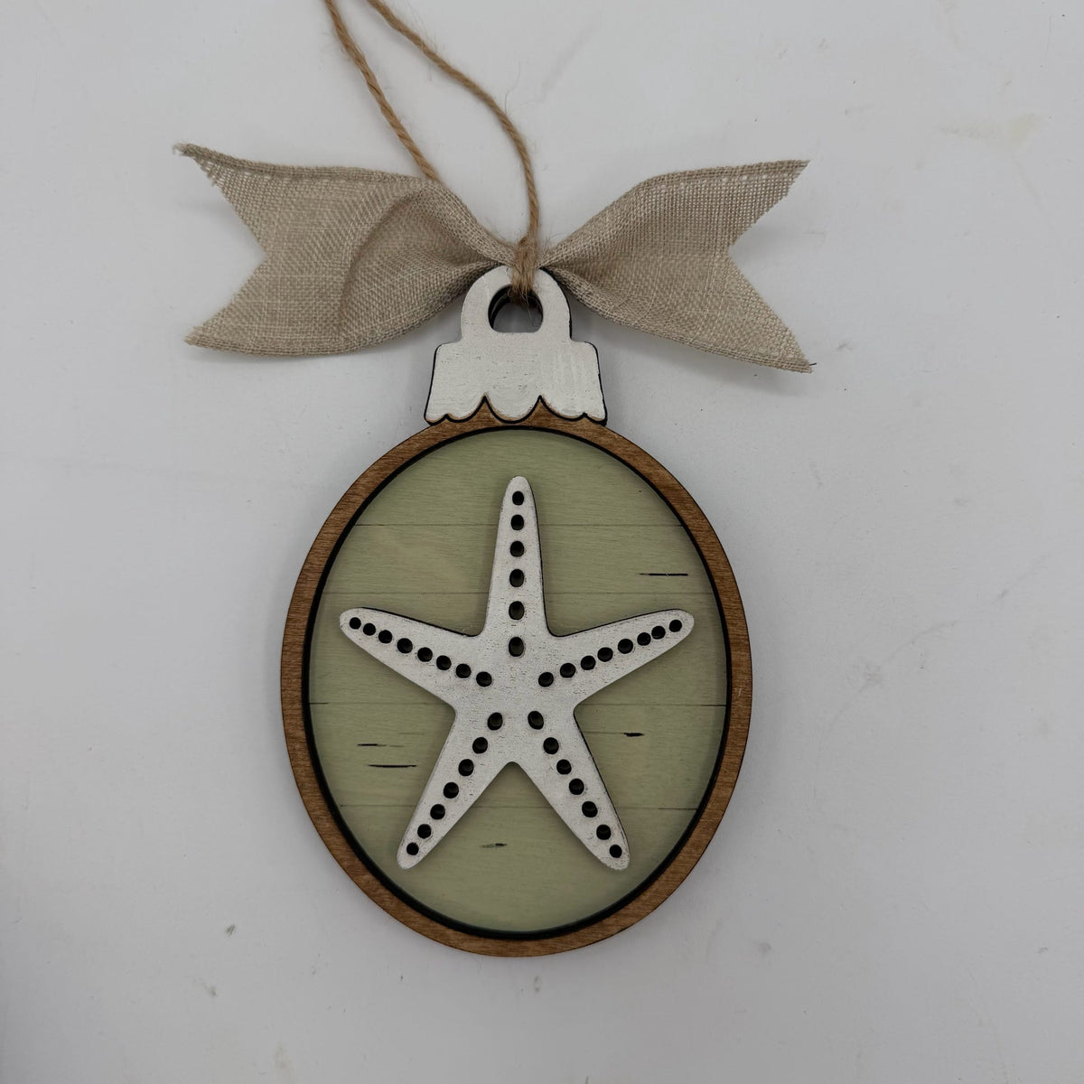 A round wooden hanging ornament with a natural wood frame, a pale green center, and a white starfish design in the middle. The ornament is suspended by a twine string and tied with a beige burlap bow at the top, shown against a plain white background.
