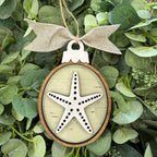 A decorative oval hanging ornament with a natural wood rim, a burlap bow at the top, and a white starfish design over a pale wood background, displayed against green foliage.
