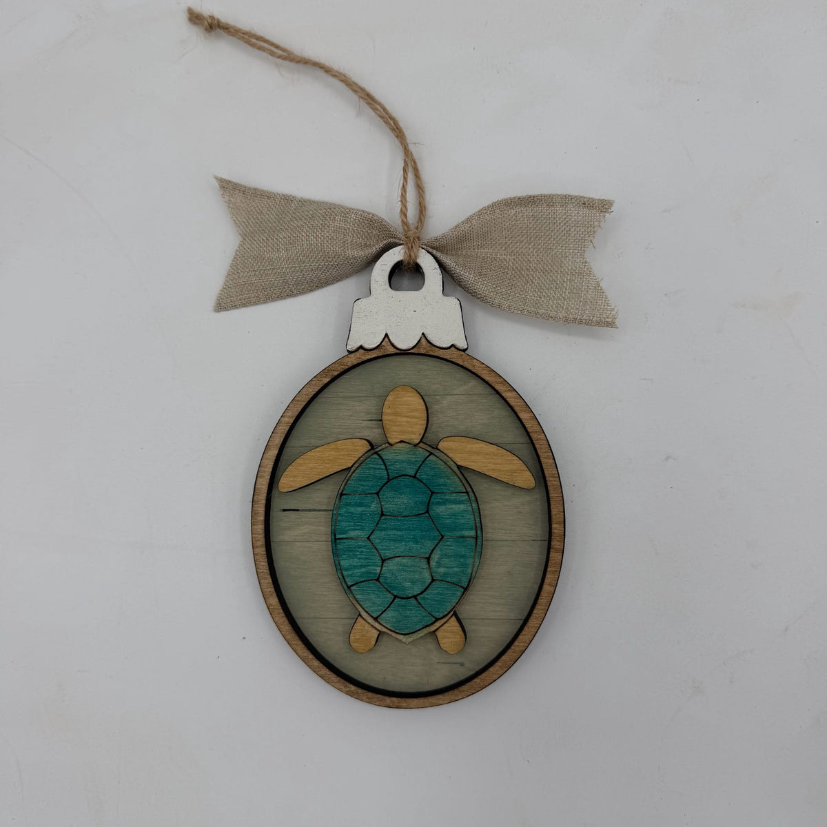 A wooden sea turtle ornament with a teal painted shell and natural wood limbs, hanging from a burlap ribbon bow on a light gray background.