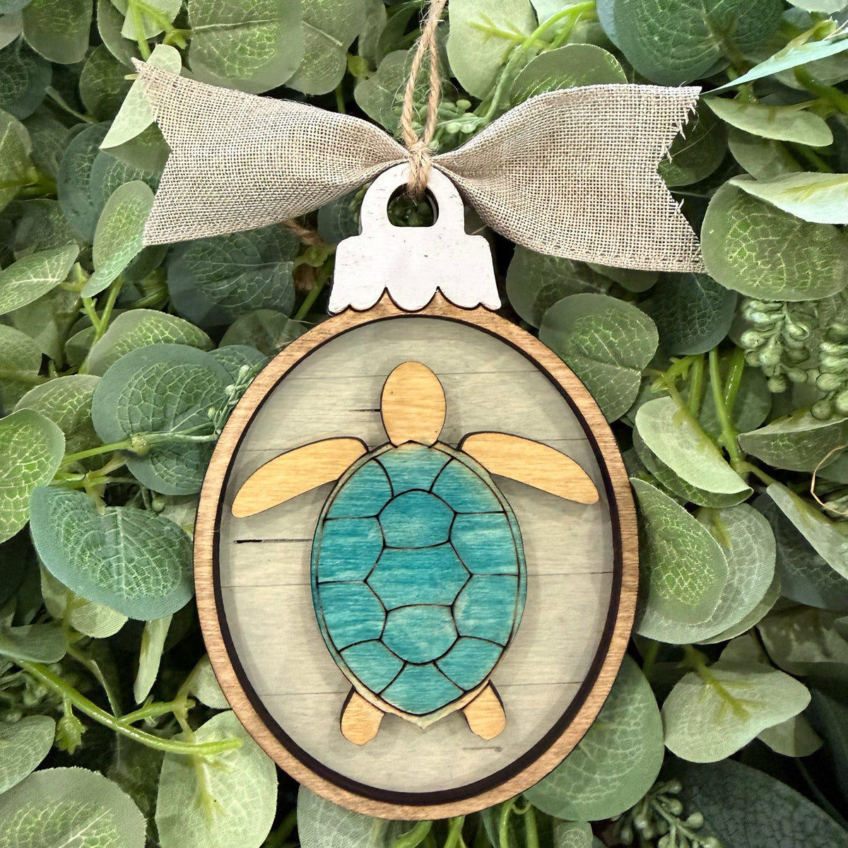 A wooden turtle ornament hanging from a burlap ribbon bow, set against a background of green eucalyptus leaves. The ornament features a light wood border, a turquoise painted shell, and natural wood accents on the flippers and legs.