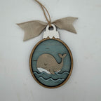 A wooden whale-shaped hanging ornament with a blue wood-grain background, layered whale and wave cutouts, a natural wood border, and a beige burlap bow on a twine loop, shown against a light gray background.