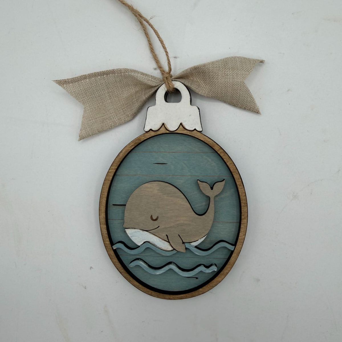 A wooden whale-shaped hanging ornament with a blue wood-grain background, layered whale and wave cutouts, a natural wood border, and a beige burlap bow on a twine loop, shown against a light gray background.