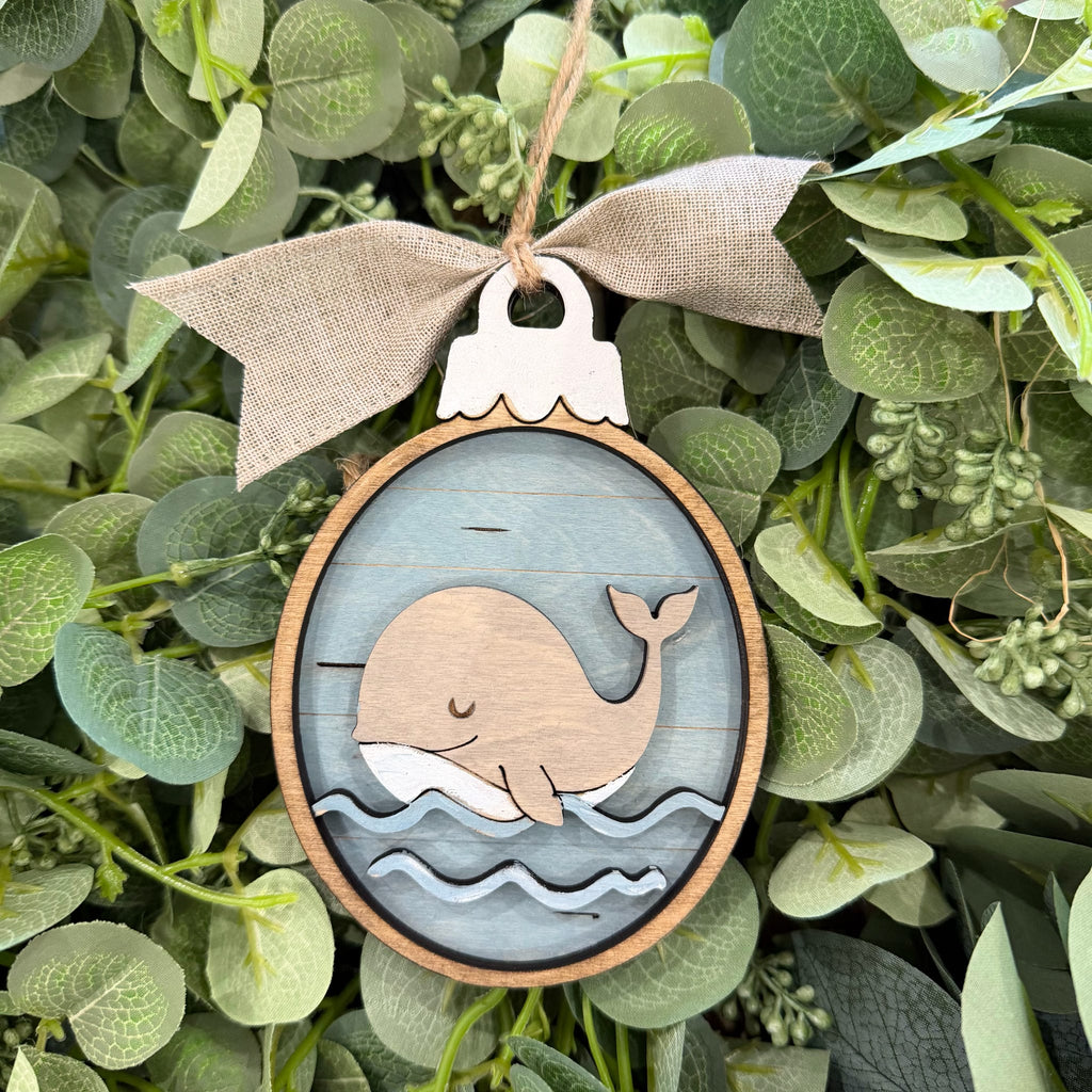 A round wooden whale ornament hanging from a beige burlap ribbon bow, set against a background of green eucalyptus leaves. The ornament features a layered wood design with a light wood whale on a weathered blue-gray wood background with white waves.