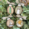 Four oval wooden Christmas ornaments with burlap bows, featuring nautical and holiday designs including a surfboard, lighthouse, sailboat, and Santa hat, displayed on green foliage.