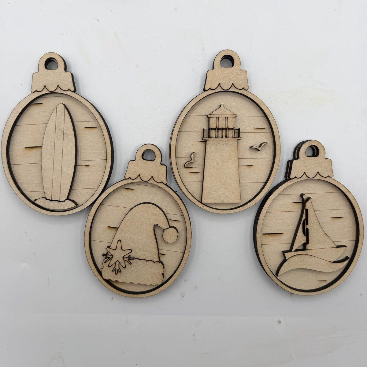 Four oval laser-cut wooden ornaments with ocean-themed designs including a surfboard, lighthouse, wave, and sailboat, displayed on a white background.