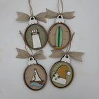 Four oval wooden hanging ornaments with burlap ribbon bows, each featuring a different nautical or coastal design: a lighthouse, a surfboard, a sailboat, and a Santa hat on a light background.