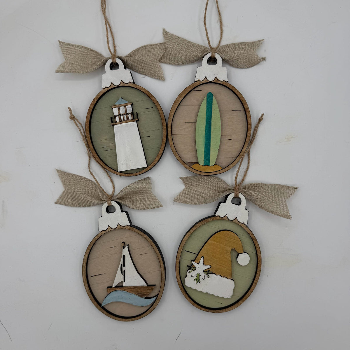 Four oval wooden hanging ornaments with burlap ribbon bows, each featuring a different nautical or coastal design: a lighthouse, a surfboard, a sailboat, and a Santa hat on a light background.