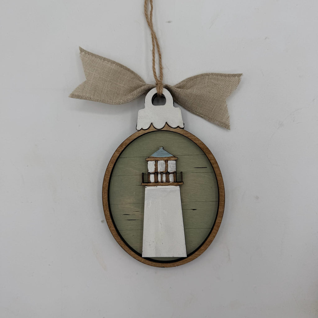 A rustic lighthouse-shaped wooden hanging ornament with a light green oval background, a white lighthouse figure, a natural wood border, and a beige burlap bow on a twine hanger against a plain light gray wall.