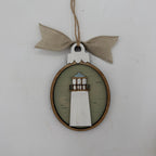 A rustic lighthouse-shaped wooden hanging ornament with a light green oval background, a white lighthouse figure, a natural wood border, and a beige burlap bow on a twine hanger against a plain light gray wall.