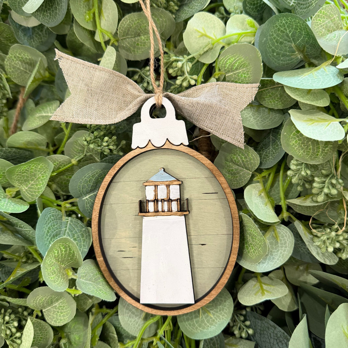 A decorative oval wooden ornament featuring a white lighthouse design with a light wood border, hanging from a beige burlap ribbon bow against a background of green eucalyptus leaves.