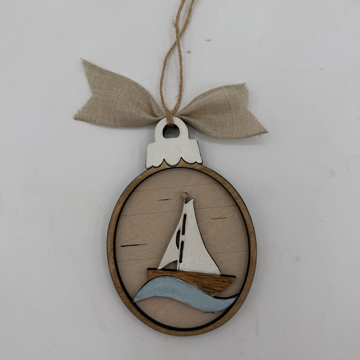 A wooden oval hanging ornament featuring a sailboat design with a light wood sail, brown hull, and light blue wave, suspended from a beige burlap ribbon on a white background.