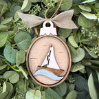 A wooden oval hanging ornament featuring a laser-cut sailboat design with a white sail, brown hull, and light blue waves, suspended from a burlap ribbon bow and placed among green leaves.