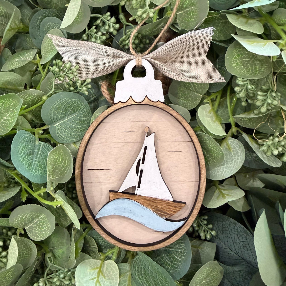 A wooden oval hanging ornament featuring a laser-cut sailboat design with a white sail, brown hull, and light blue waves, suspended from a burlap ribbon bow and placed among green leaves.