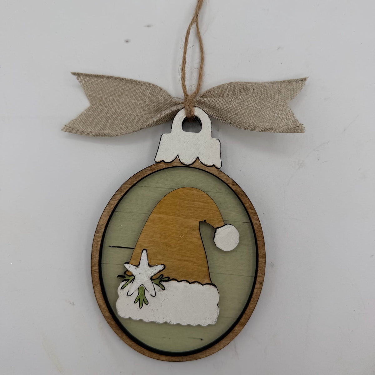 A round wooden Christmas ornament featuring a layered design with a brown Santa hat, white faux fur trim, and a light green background, hanging from a beige burlap ribbon bow.