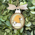 A round wooden Christmas ornament featuring a layered Santa hat design in natural wood and white, hanging from a beige burlap ribbon bow against a background of green eucalyptus leaves.