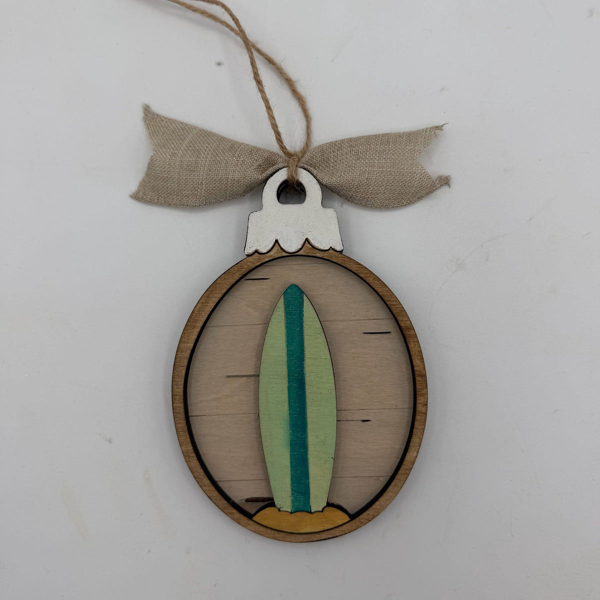 A wooden oval hanging ornament featuring a painted surfboard design in light green, teal, and tan, suspended from a burlap ribbon on a plain white background.