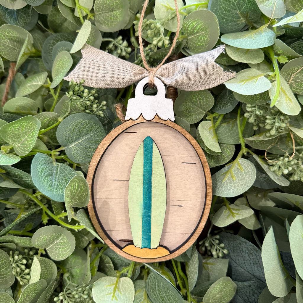 A wooden oval ornament featuring a layered surfboard design in light green, teal, and tan, hanging from a burlap ribbon against a background of green eucalyptus leaves.