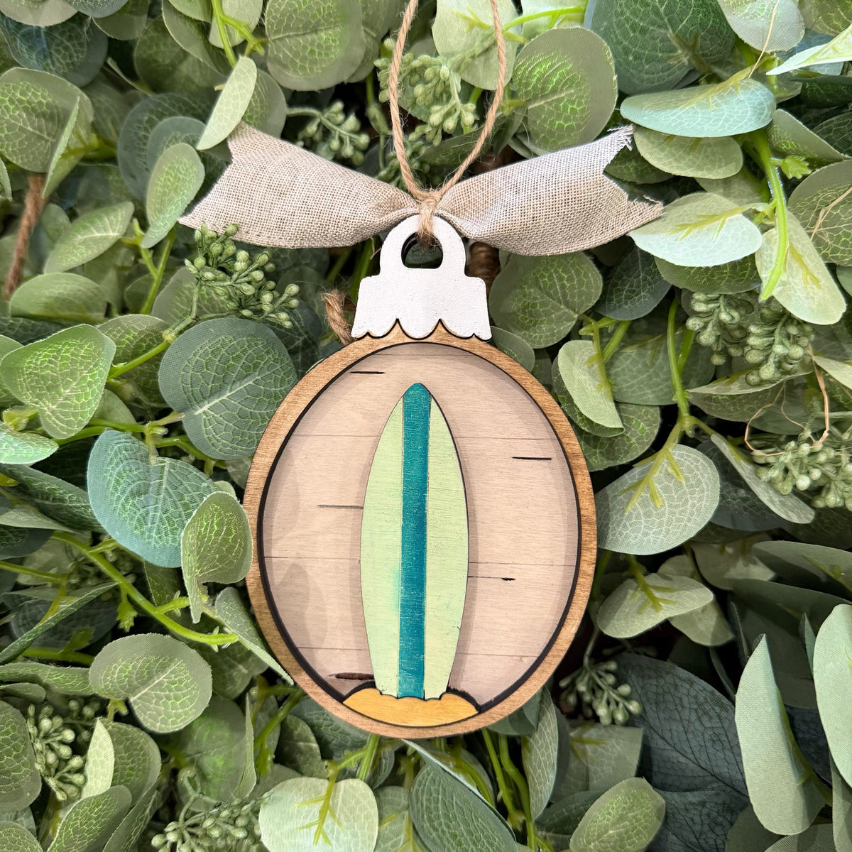 A wooden oval ornament featuring a layered surfboard design in light green, teal, and tan, hanging from a burlap ribbon against a background of green eucalyptus leaves.