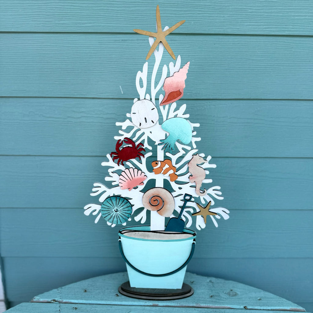 A decorative metal coastal tree centerpiece with seashells, a starfish topper, and a small bucket base, displayed against a teal wood plank wall.