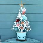 A decorative metal coastal tree centerpiece with seashells, a starfish topper, and a small bucket base, displayed against a teal wood plank wall.