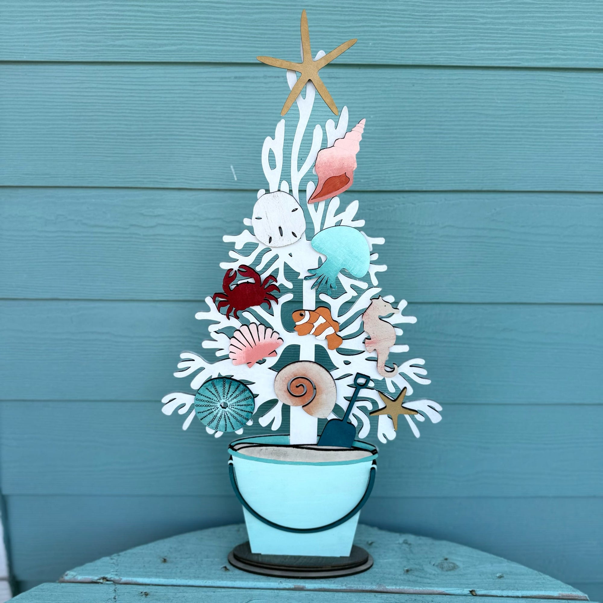 A decorative metal coastal tree centerpiece with seashells, a starfish topper, and a small bucket base, displayed against a teal wood plank wall.