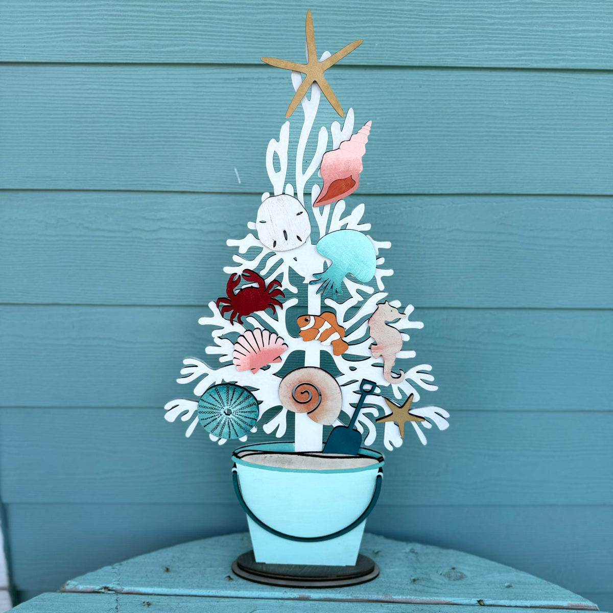A decorative metal coastal tree centerpiece with seashells, a starfish topper, and a small bucket base, displayed against a teal wood plank wall.