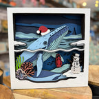 A square layered paper-cut Christmas shadow box artwork featuring a blue whale wearing a red Santa hat, a snowman, a red lantern, coral, and snowy mountains, displayed on a wooden surface with a blurred shop background.