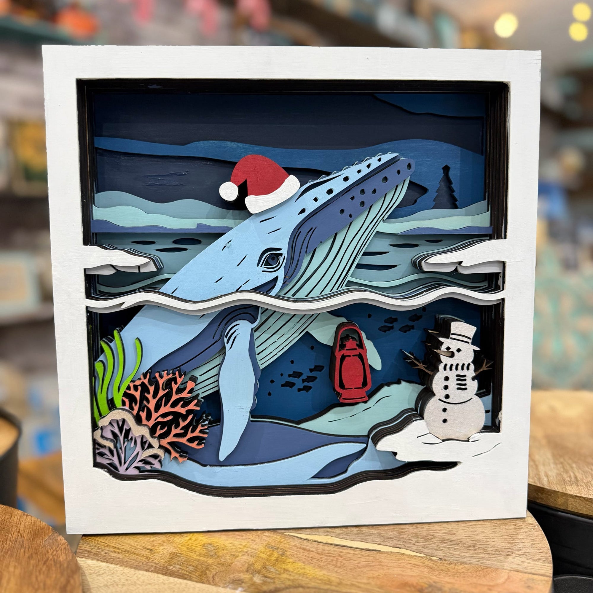 A square layered paper-cut Christmas shadow box artwork featuring a blue whale wearing a red Santa hat, a snowman, a red lantern, coral, and snowy mountains, displayed on a wooden surface with a blurred shop background.