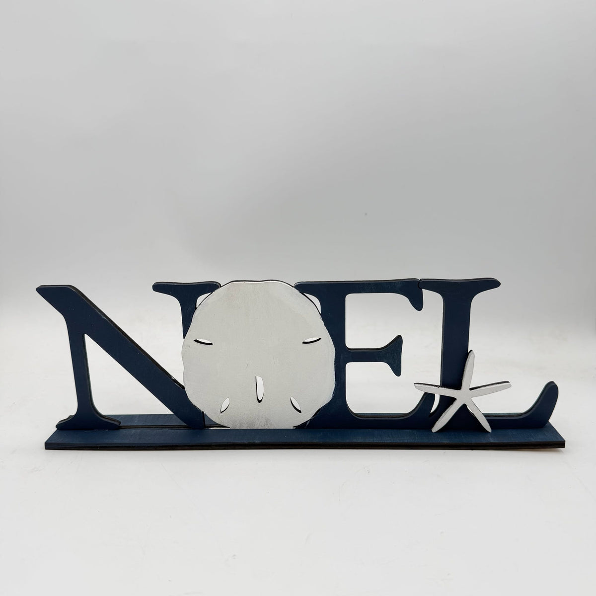A nautical-themed decorative sign spelling 'NOEL' in dark blue, featuring a silver sand dollar in the center and a small silver starfish on the right, set against a neutral gray background.