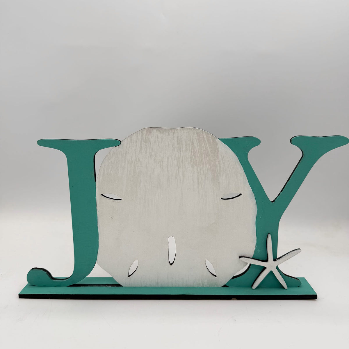 A decorative wooden sign spelling 'JOY' with a large seashell in the center, teal letters on a base, and a small starfish accent on the right, photographed against a plain light gray background.