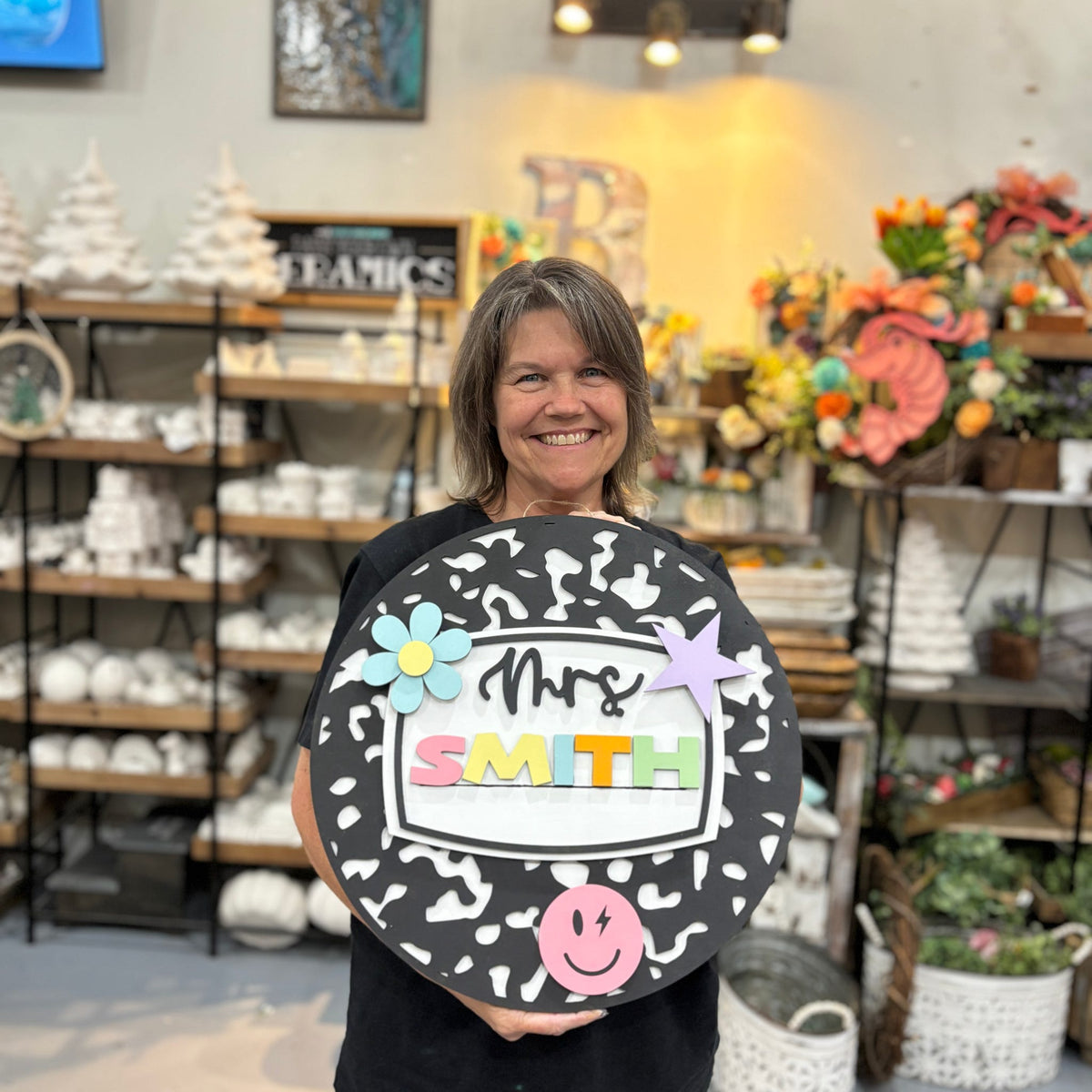 A smiling woman stands in a craft or home decor store holding a large round black sign with white irregular spots. The sign reads "Mrs.