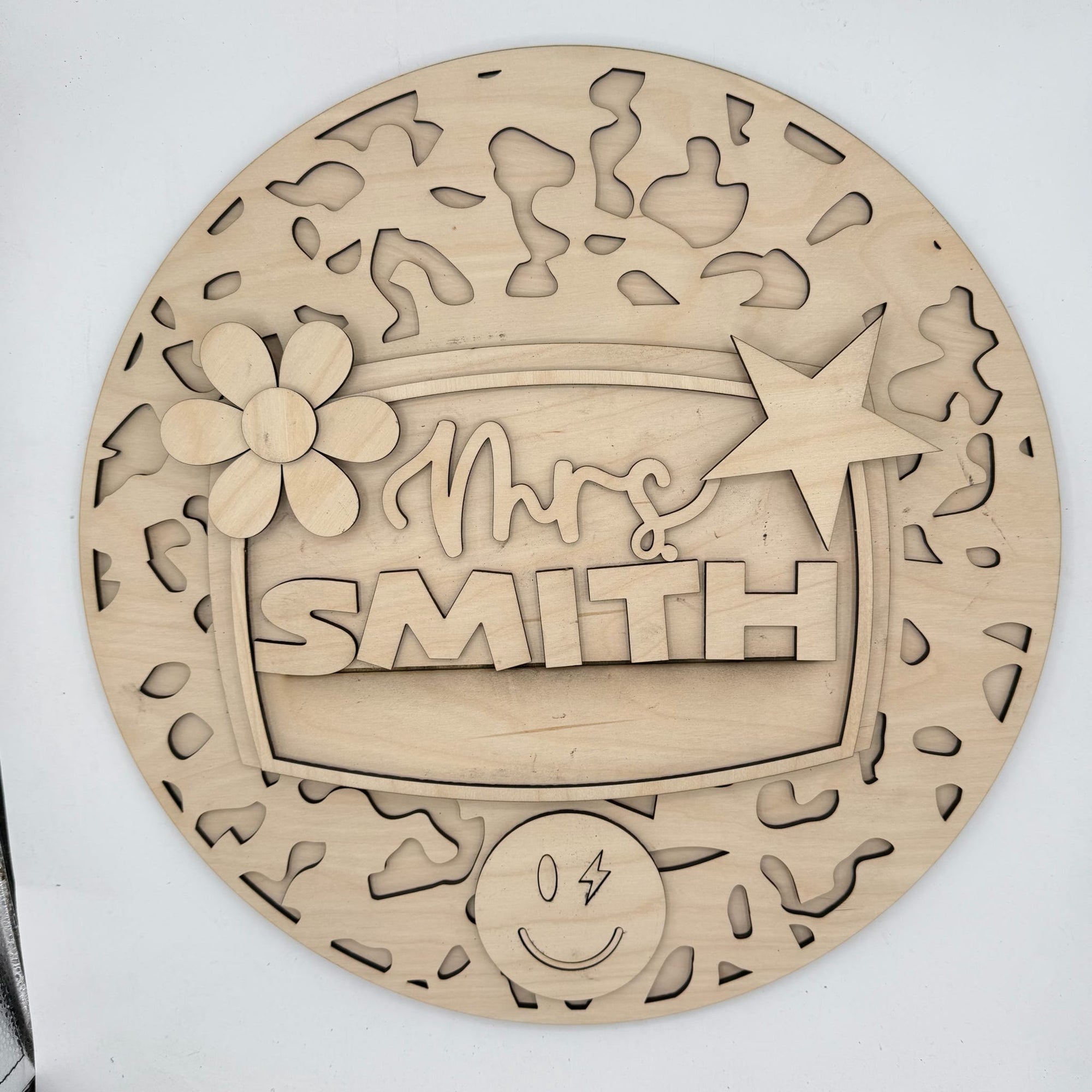 A round, laser-cut wooden plaque or sign with cutout patterns and raised text reading 'Mrs. SMITH,' plus decorative floral and star details, shown on a plain light background.