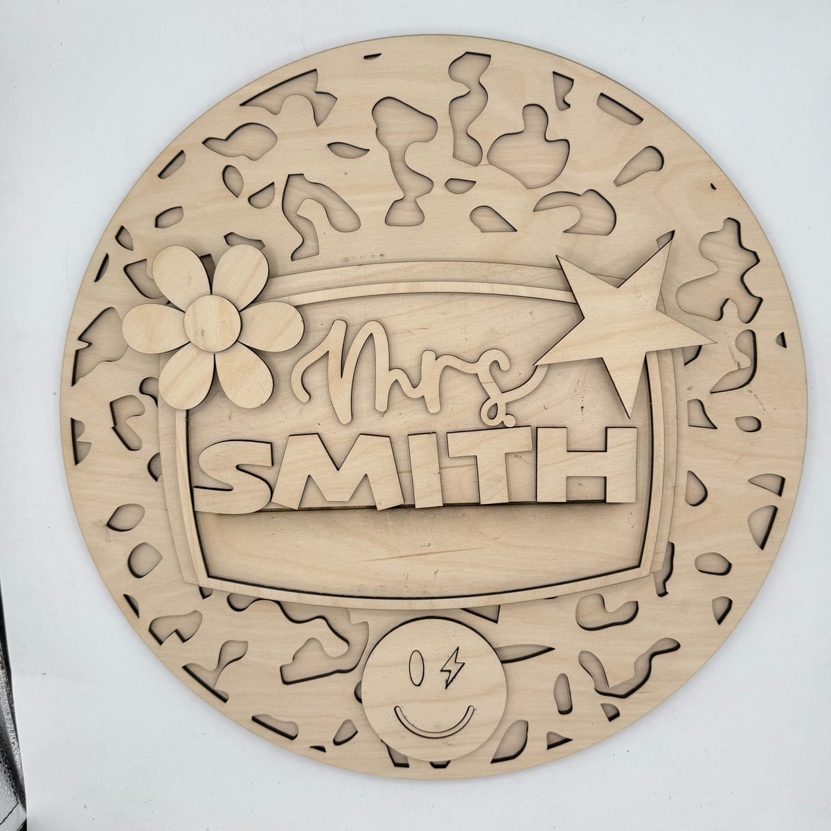 A round, laser-cut wooden plaque or sign with cutout patterns and raised text reading 'Mrs. SMITH,' plus decorative floral and star details, shown on a plain light background.