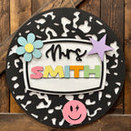 A personalized round black wooden wall sign with a white center panel reading 'Mrs. SMITH' in colorful letters, decorated with a blue flower, purple star, pink smiley face, and a pink nameplate, shown against a rustic wooden plank background.