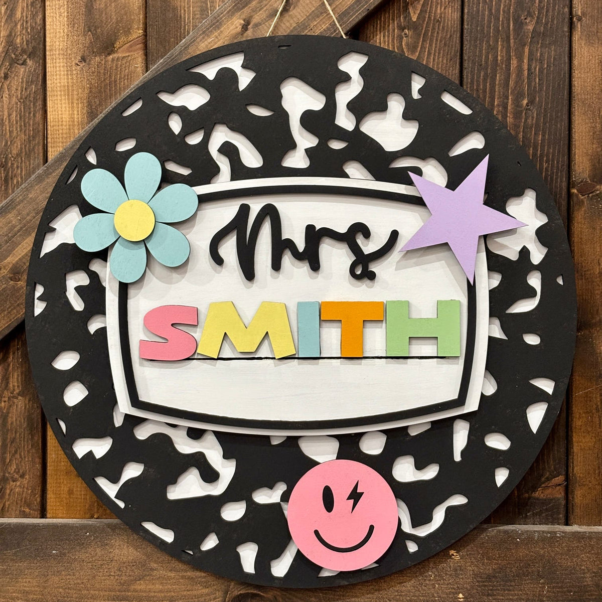 A personalized round black wooden wall sign with a white center panel reading 'Mrs. SMITH' in colorful letters, decorated with a blue flower, purple star, pink smiley face, and a pink nameplate, shown against a rustic wooden plank background.
