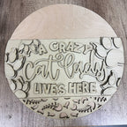 Round wooden sign with 'A crazy cat lady lives here' text and cat illustrations on a wooden surface.