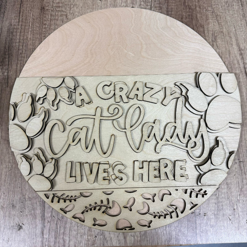 Round wooden sign with 'A crazy cat lady lives here' text and cat illustrations on a wooden surface.