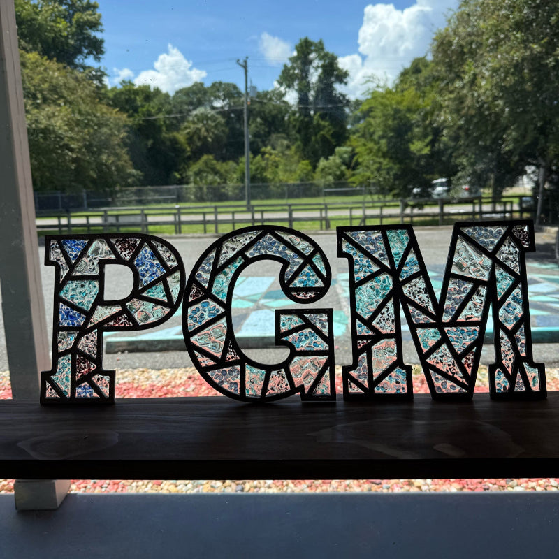 Decorative letters 'PGM' with a mosaic pattern in front of a window with a view of trees and a road.