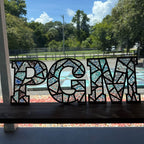Decorative letters 'PGM' with a mosaic pattern in front of a window with a view of trees and a road.