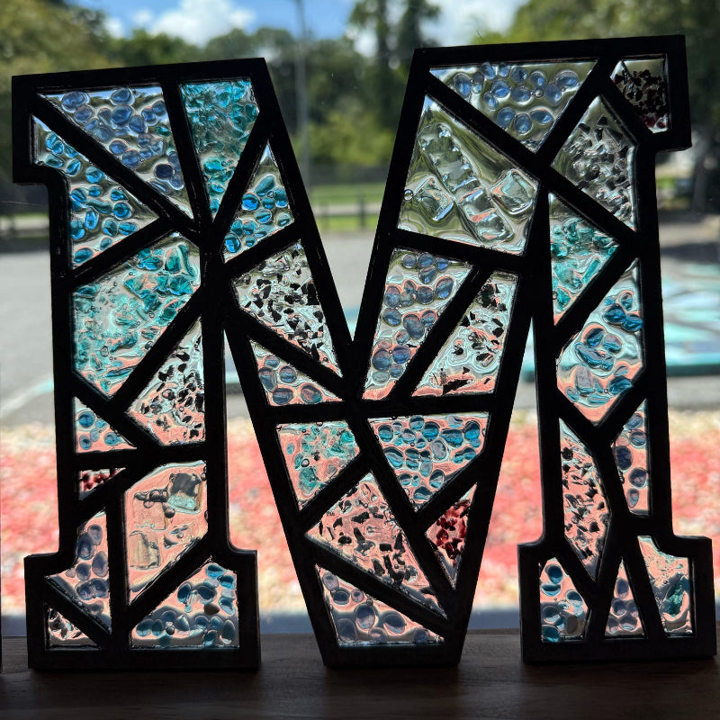Decorative letter 'M' with a mosaic design in front of a window.