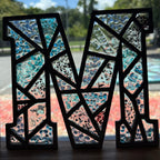 Decorative letter 'M' with a mosaic design in front of a window.