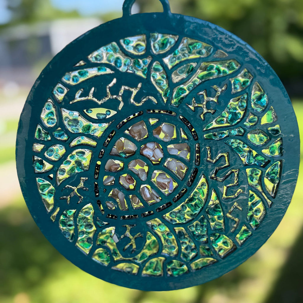A circular stained glass suncatcher with a teal painted metal frame and intricate cutout floral patterns, featuring iridescent green and gold glass panels, shown outdoors against a blurred green background.