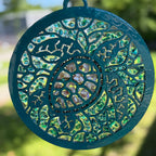A circular stained glass suncatcher with a teal painted metal frame and intricate cutout floral patterns, featuring iridescent green and gold glass panels, shown outdoors against a blurred green background.