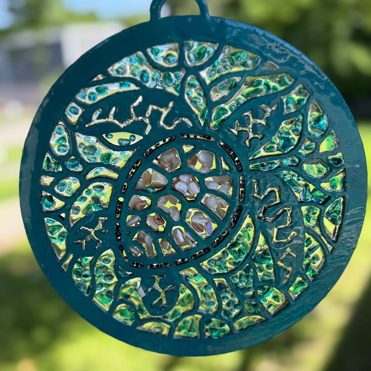 A circular stained glass suncatcher with a teal painted metal frame and intricate cutout floral patterns, featuring iridescent green and gold glass panels, shown outdoors against a blurred green background.