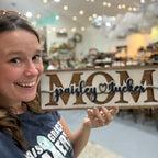 Woman holding a sign that reads 'MOM' with a heart and text in a store setting.