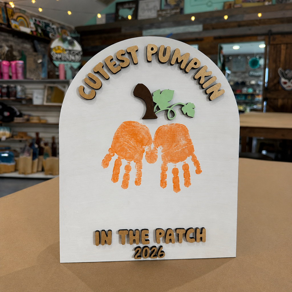 A white arched plaque or sign with laser-cut wooden text reading 'CUTEST PUMPKIN' at the top and 'IN THE PATCH 2026' at the bottom, featuring orange handprint pumpkin shapes and a small green leaf sprig in the center. The sign is photographed on a tabletop inside a store with shelves and decor blurred in the background.