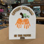 A white arched plaque or sign with laser-cut wooden text reading 'CUTEST PUMPKIN' at the top and 'IN THE PATCH 2026' at the bottom, featuring orange handprint pumpkin shapes and a small green leaf sprig in the center. The sign is photographed on a tabletop inside a store with shelves and decor blurred in the background.