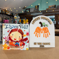 Two autumn-themed products displayed side-by-side on a wooden table in a shop: a children's board book titled 'I Love Fall!' and a personalized wooden sign that reads 'CUTEST PUMPKIN IN THE PATCH 2026' with orange handprint pumpkin designs.